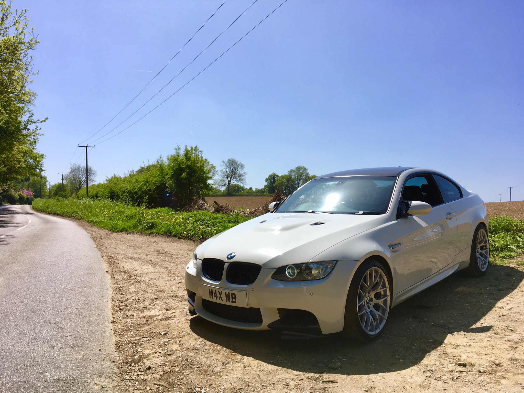 A car is parked on the side of the road - Pistonheads - The image features a silver BMW car parked on the side of a road. The vehicle is in good condition and appears to be in motion, as suggested by its dynamic position. The surroundings include a rural landscape with a dirt path leading towards some greenery in the distance. The sky is partly cloudy, indicating fair weather conditions. There are no visible texts or distinctive marks that would provide additional context or information.