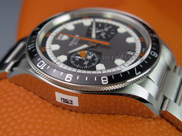 Incoming..what do you have? (Vol. 3) - Page 260 - Watches - PistonHeads - The image shows a close-up of a wristwatch with a silvered metal band and buckle, positioned on an orange textured surface which appears to be an article of clothing or a watch strap. The watch face is black with white and orange accents, featuring a tachymeter scale on the outer edge. The second hand is prominent against the black background. The brand name is visible on the dial in silver letters, while the bezel is adorned with distinct orange markers. There is a small, rectangular tag with text at the bottom of the image, but the text is not clearly legible.