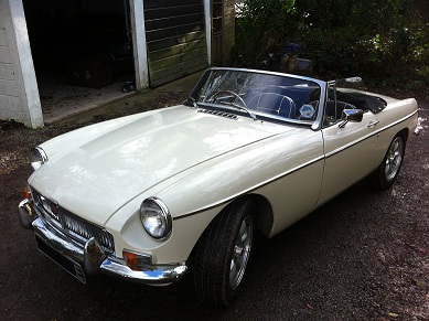 MGB buying tips - Page 1 - MG - PistonHeads - The image shows a cream-colored, classic sports car parked on a surface that appears to be made of gravel or fine rock. The vehicle has a convertible top up and is styled with a clean and sleek aesthetic. The car is equipped with a pair of circular headlights and has a round, emblematic logo on the grille, which is a distinctive feature of many classic models. The car is angled slightly to the left of the frame, and the surrounding environment suggests a rural or semi-rural setting with greenery in the background.