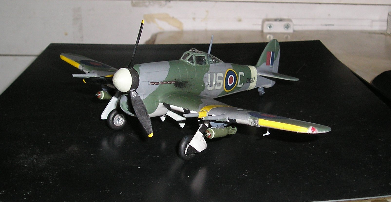 Frog Rovex Hawker Pistonheads Typhoon - This image depicts a miniature model plane on a surface that appears to be a factory floor. The plane is a vintage design, predominantly camouflaged in shades of green and grey, with white and yellow markings. It has a single propeller and is shown from a side angle. The cockpit can be seen on the top of the plane with the letters "US" visible. There is a reflection of light on the surface below the model plane, and the setting suggests an indoor environment, possibly a workshop or assembly line.