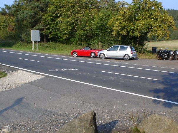 Slow Mark Pistonheads Feel Clio - The image depicts a serene scene of a country road near a wooded area. Two cars, one red and one silver, are parked on the side of the road, possibly at a rest stop or simply pulled over. The road is lined with trees on both sides, suggesting a rural or semi-rural location. The grassy area surrounding the road is verdant, indicative of a healthy, thriving environment. The overall scene conveys a sense of tranquility and relaxation.