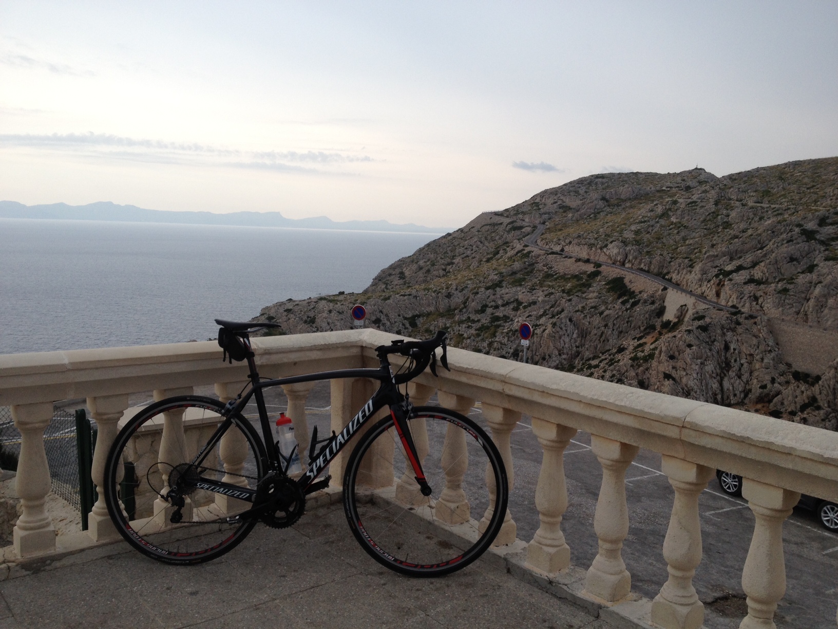 The "Photos From Today's Ride" thread... - Page 398 - Pedal Powered - PistonHeads - The image displays a stark contrast between the man-made and the natural. A sleek black racing bike is parked on a balcony, facing the water. The bike, with its distinctive red, white, and blue color scheme, is propped against a balustrade that commands a breathtaking view of the ocean. The calm blue of the sea stretches out to the horizon, where mountains rise in the distance, suggesting an adventurous ride might be in the near future. The partially cloudy sky above adds to the dramatic scenery, offering a sense of tranquility and exhilaration at the same time.