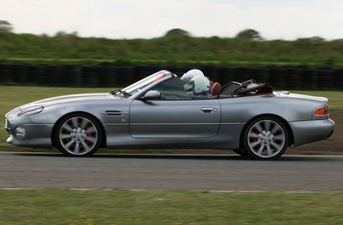 The Proper Colour for an Aston thread - silver, greys, black - Page 1 - Aston Martin - PistonHeads - The image shows a silver Aston Martin sports car in motion on a road. The car is occupied by a driver visible through the glass, with a pink or coral-colored top visible on the car, suggesting it is a convertible model. The driver appears to be wearing a helmet, indicating a focus on safety. The car is driving in front of a field and a line of trees, on an overcast day. The background shows that the road is on a track, possibly a gray or concrete surface, given the manicured appearance of the grass on either side.