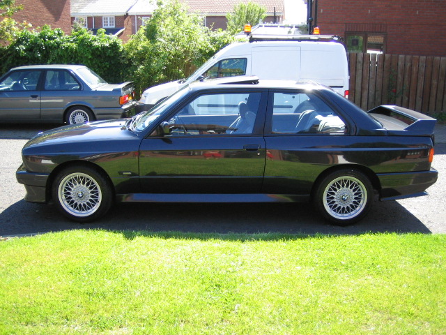 Contribution Pistonheads - This image captures a serene scene in a residential area. The main focus is a black sports car parked on the grass, adjacent to a driveway. The car is sleek and stylish, Its dark hue contrasting with the surrounding environment. The reception area of the house is visible, where a Mercedes saloon car is parked, adding an air of luxury to the setting. The backdrop of the image is composed of houses and trees, creating a peaceful suburban atmosphere.