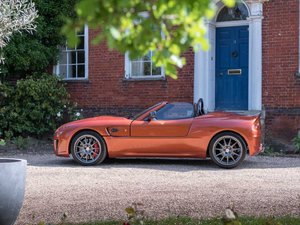 The I'm Bored Guess The Car Quiz (No Googling allowed) - Page 22 - Classic Cars and Yesterday's Heroes - PistonHeads - The image displays a vibrant red sports car parked in front of a traditional British brick building. The car is a convertible with the top down, showcasing its interior. It's equipped with silver rims and a spoiler on the back. In the background, there are trees visible through an archway, suggesting a residential area. A person appears to be in the process of taking the photograph from inside the car, adding a human element to the scene. The setting seems serene, with the focus being on the car itself and its contrasting colors against the brickwork.