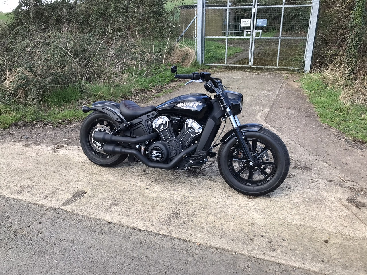 Pistonheads - The image features a black and chrome motorcycle parked on a concrete surface. The motorcycle has a prominent front fork design, with the engine clearly visible beneath the fuel tank. It is parked in an outdoor area, possibly near a road or pathway, as suggested by a fence and green foliage in the background. There is no text present in the image.
