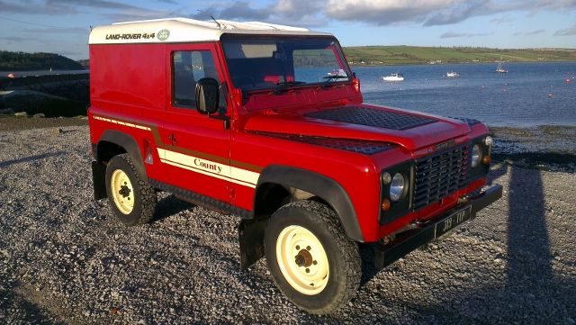 Here is my new toy - Page 1 - Ireland - PistonHeads - The image captures a red Land Rover 90, standing confidently on a gravel surface. The vehicle is equipped with a white roof rack, suggesting a readiness for adventure or simply storage. Behind it, there's a tranquil scene of a body of water dotted with buoys and boats, creating a serene backdrop to the rugged vehicle. The blue sky and the presence of a single person at some distance add a touch of life to the otherwise inanimate scene.