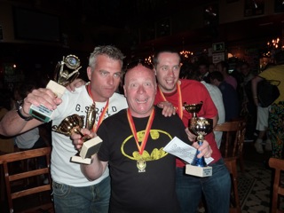 "Chip run cars win in Barcelona" - Page 1 - East Anglia - PistonHeads - This image captures a joyful moment of celebration and camaraderie. There are three men standing close to each other, each holding a distinctive trophy. They are the main subjects in the image. The man on the left is holding a trophy that appears to have some sort of ribbon or sash. The center man, wearing a Batman T-shirt, is holding his trophy along with a green object, possibly a certificate or award. The man on the right is holding a trophy that includes both a cup and a medal, symbolizing multiple awards.

In the background, there are other people, possibly organisers or fellow competitors, enhancing the feel of an event or competition. The setting seems to be indoors, possibly a banquet hall or a large room, given the blurred backdrop and the furniture visible such as chairs and a dining table.

The key focus of the image is the interaction between the three men, with their triumphant poses against the backdrop of others, perhaps looking on with admiration or anticipation. The image conveys a sense of achievement and sportsmanship, capturing a moment of personal triumph in the context of a collective event.