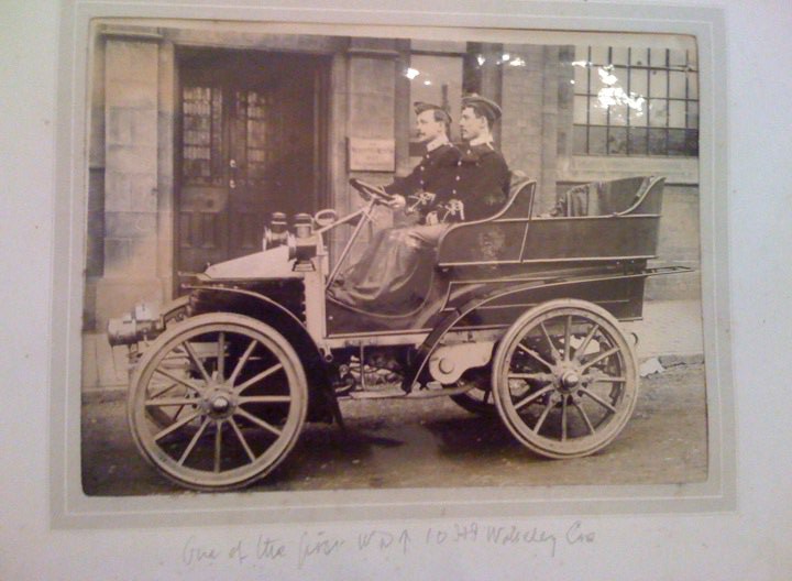 Wolesley Century Pistonheads Turn - The image is a black and white photograph capturing a quaint moment in time. It features a horse-drawn carriage parked on a street. Within the carriage, there are two individuals, likely a driver and a passenger, both dressed in clothing indicative of an earlier era. The driver is seated on the left side of the carriage, and the passenger is seated to the right. The carriage is adorned with a coat of arms, adding a touch of nobility or importance to its function. The background reveals a building and some upper windows, typical of urban architecture from the past. The image is framed and possibly carries an inscription at its bottom, though it's not fully visible. The entire scene is a glimpse into a bygone era, offering a window into historical transportation modes.