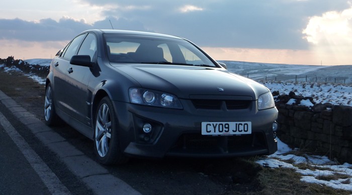 My VXR8 - pics taken today - Page 1 - Readers' Cars - PistonHeads - The image presents a striking scene set against a backdrop of a winter landscape. Dominating the foreground is a black car, parked by a stone wall on the right side of a road. The car, which is a Mazda CX-9, is facing the camera, its headlights and curved front grille adding to the overall aesthetic. In the distance, a mountain range stretches across the horizon, their peaks blanketed in patches of snow. Above, the sky is filled with clouds, hinting at an overcast day. The overall scene paints a picture of a tranquil, snowy day in a mountainous region.