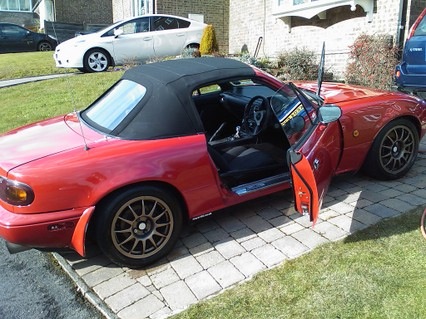 Pistonheads Megamile - The image showcases a vibrant red sports car, parked on a brick driveway in front of a house. The car's top is down, inviting you to take in the interior, which includes a black interior and black seat covers. The bright sunlight of the day matches the car's color, casting appealing shadows and creating a dynamic visual scene. In the background, there's a hint of a blue car, but it's clearly overshadowed by the striking red sports car. Each element in the scene contributes to a sense of leisure and the joy of driving.