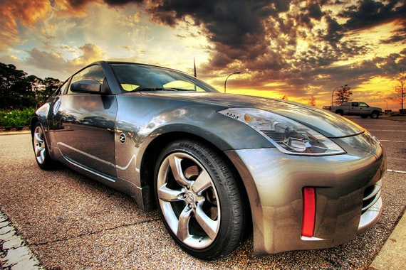 Some half decent pics - Page 1 - North West - PistonHeads - The image displays a modern silver car parked on asphalt. The car features a sleek and aerodynamic design, with a distinctive grille and a smooth body. The wheels are silver and have a multi-spoke alloy design. The background of the image is characterized by a dramatic sunset or sunrise with clouds in the sky, which suggests the photo was taken around the time of day when the sun is low on the horizon. The colors in the sky range from orange to yellow, and there are a few trees visible in the upper part of the image, indicating the car is parked in an open area.