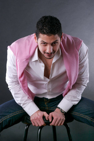 The image showcases a man with a relaxed posture, donning a pink vest and a white shirt. He stands with his arm perched on the armrest of a chair. His facial expression is serious, adding a touch of intensity to the otherwise casual image. The photograph captures the man against a neutral background, ensuring that the focus remains on him.