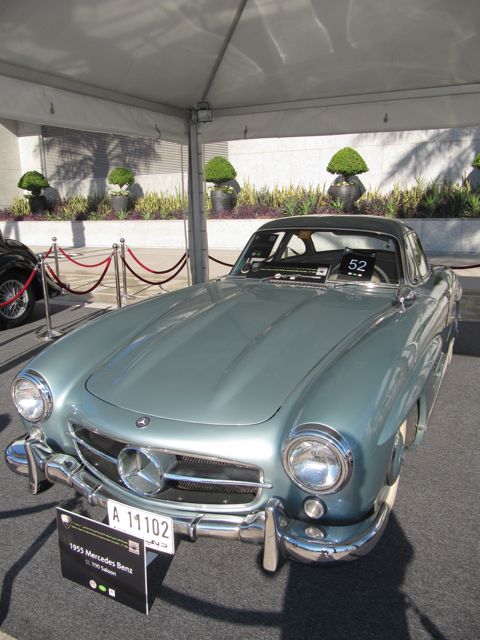 Pistonheads Emaar Classic - In the foreground, there's a vintage Mercedes Benz car on display at a car show. The car, with its silver metal body and black interior, radiates an aura of elegance and luxury. Its registration number, A1102, is prominently displayed on the front amidst a variety of other car-related information. The car is positioned under a white canopy, which provides shelter and underscores the event's professionalism. The tarmac where it's parked fades into a blur in the background, drawing the viewer's focus on the beautifully maintained vintage vehicle.