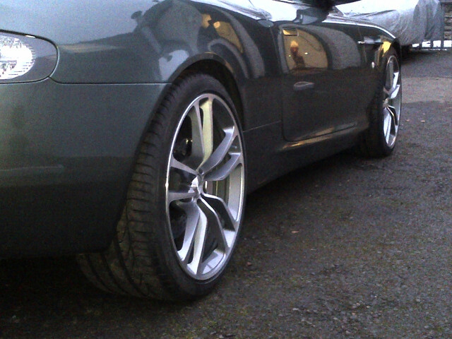 DBS wheels on DB9 - (more or less) - Page 1 - Aston Martin - PistonHeads - The image showcases the front of a dark-colored car parked on a paved road. The vehicle is set against the backdrop of a clear sky, highlighting its reflective surface. A distinctive feature of the car's front is its large, black-rimmed wheels that contrast with the shiny car body. The car appears to be in good condition, reflecting the care and maintenance it receives. Its parked position suggests that the owner has momentarily stepped away and will return shortly.