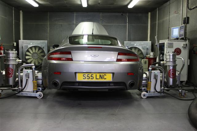 V8 Vantage tuning (SLINC) - TDI Inspection Service - Page 1 - Aston Martin - PistonHeads - This image depicts a scene within an auto repair facility. A silver car, bearing the license plate "SS5 LNC," is parked inside, next to what appears to be an exhaust system or a device for mitigating vehicle emissions. There are signs of maintenance or diagnostic work, as evidenced by the multiple equipment pieces surrounding the vehicle. A few filters, possibly related to the car's exhaust system, are visible beside the rear tire. The environment suggests a professional and technical setting focused on automotive care.