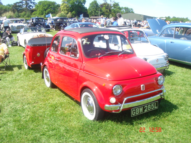 Old Warden car show - Page 1 - Classic Cars and Yesterday's Heroes - PistonHeads - The image showcases a field of vibrant colors where several antique cars are parked side by side, exhibiting a variety of shades, including red, blue, and white. The cars are arranged neatly, and a small traffic light is situated in the center of the lineup. A crowd of people is seen admiring the cars in the background, suggesting a car show or similar event. The scene radiates a relaxed and nostalgic atmosphere, indicative of a leisurely and enjoyable day out.