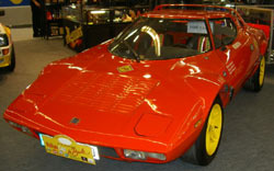 Beautiful Pistonheads Bertones - The image displays a vintage sports car with an orange body and a yellow interior. The car is parked indoors, shown from a three-quarter angle, giving a good look at its front and the passenger side. It has a curved nose and sits on yellow-trimmed wheels. The vehicle appears to be the main focus of the image in a display setup, and there are other vintage cars in the background. A solitary figure can be seen in the far left background, but they are not interacting with the car.