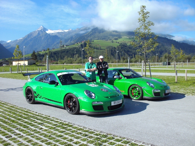 Viper Green 997 GT3RS - Page 4 - Porsche General - PistonHeads - The image captures a serene scene in a parking lot in the mountains. Two vibrant green Porsche cars are the focal point, parked side by side. Standing proudly next to them are two men, donned in white helmets and clad in green shirts, perhaps the proud owners of these sleek vehicles. The backdrop features majestic mountains under a clear blue sky, adding to the picturesque scene. The vast expanse of the parking lot stretches ahead of the cars, inviting sneak peeks of more Porsches parked farther away.