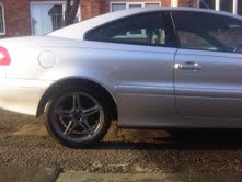 Pistonheads - The image presents a close-up view of the back corner of a silver-colored sedan parked on a street. The car's back right wheel is prominently visible in the foreground, displaying a sleek wheel design. The view is from a lower angle, which accentuates the shape of the car and gives a clear view of the wheel and its intricate details. In the background, part of a brick building can be seen, suggesting an urban setting.