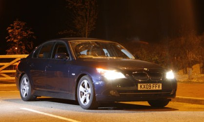 Pistonheads Unmarked - The image captures a moment on a dark street where a blue BMW car is the primary subject. The car is facing towards the right side of the image and seems to be in motion. The lights of the car are on, illuminating the car and creating a contrast with the darkness of the street. The street itself is not well-lit and is mostly shrouded in darkness, suggesting it might be late at night. In the background, there's a fence, adding to the atmosphere of solitude and travel. The image conveys a sense of quiet intensity, as if the car is on a searching journey through the night.