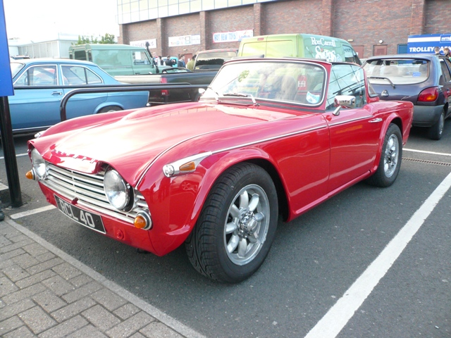 Metrocentre Pistonheads - The image showcases a vibrant red classic convertible car parked in a lot near various other vehicles. The car's polished top and chrome accents shine in the light, reflecting its past charm despite its age. It's nestled amidst other vehicles, hinting at a busy urban environment. The car's position, slightly angled towards us, invites a closer look at its details.