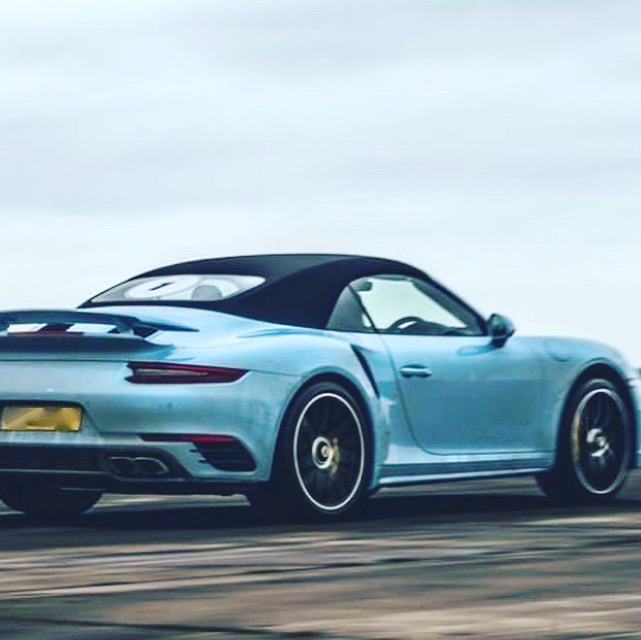 Pistonheads - The image shows a blue Porsche sports car in motion on what appears to be an empty road. The vehicle is captured from the rear, giving prominence to its aerodynamic design and the distinctive Porsche logo on the back wing. It's equipped with a large spoiler that stands out against the blue bodywork. There are two visible license plates, one at the front and another at the rear of the car. The setting suggests an overcast day due to the gray sky, which contrasts with the vibrant blue of the Porsche. The speed of the car is indicated by the motion blur on the background, suggesting it's moving quickly along the road.
