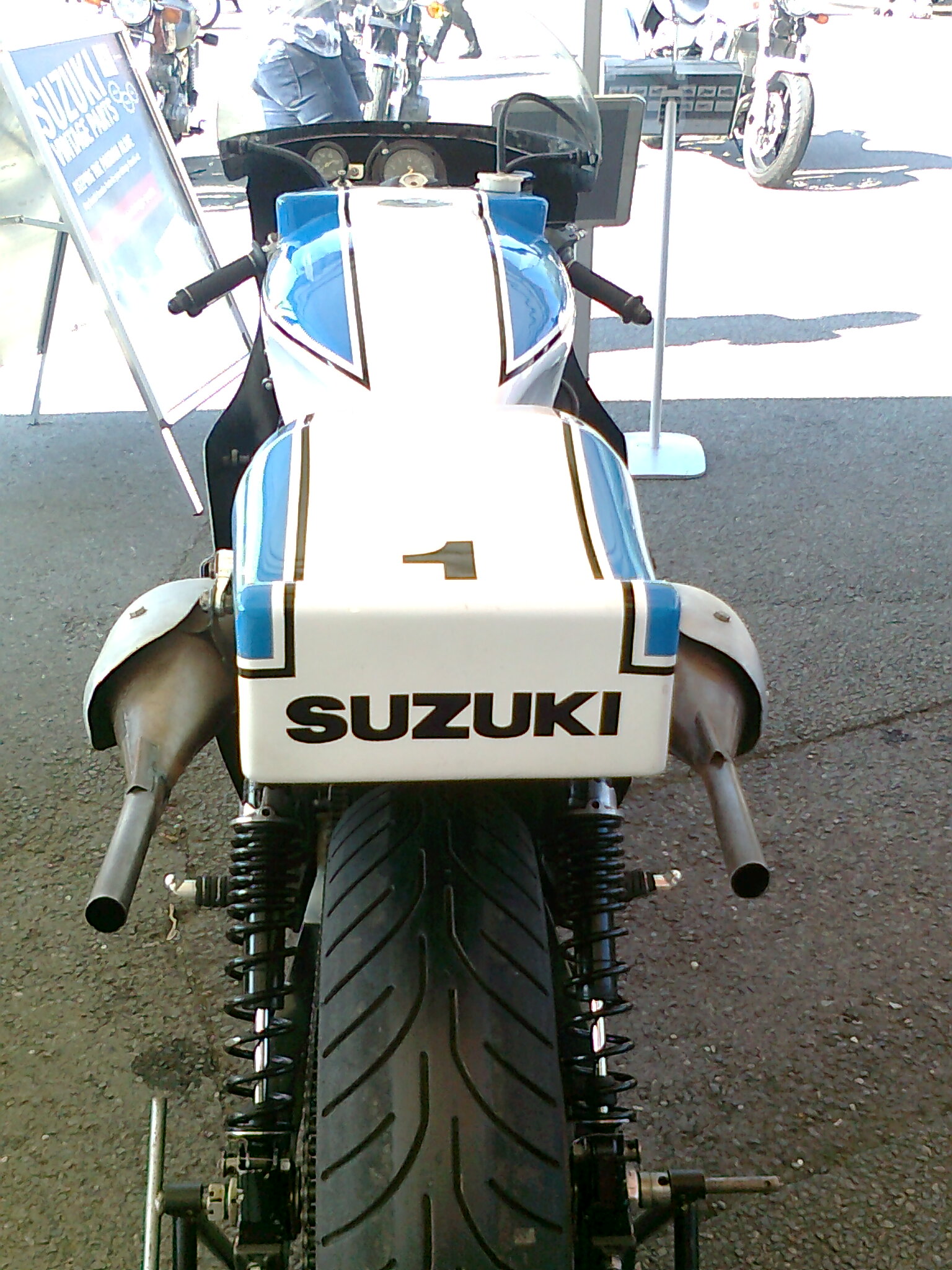 A motorcycle parked on the side of a road - Pistonheads - This image features a close-up view of the back end of a blue and white Suzuki motorcycle. The motorcycle is parked on a paved surface and its most prominent features include its rear tire, exhaust pipes, and white fending. Behind the motorcycle, there are other vehicles visible, suggesting that this might be a parking lot or showroom area. The focus of the image is clearly on the motorcycle, particularly the number one positioned on the fender, indicating it may be a racing bike or part of a series.