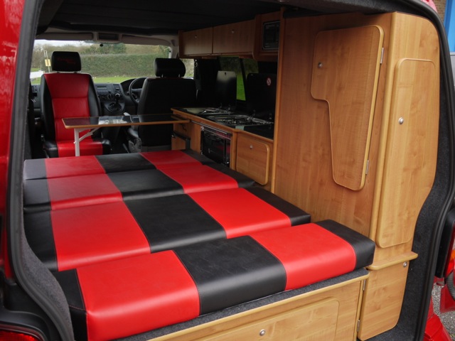 First T5 Camper - Page 1 - Tents, Caravans & Motorhomes - PistonHeads - The image depicts the interior of a van, which has been creatively modified. The main feature is a large red bench seat that covers much of the floor area, providing seating that also doubles as a bed with a black and red striped top. On the opposite side of the van is a functional kitchen setup, complete with a sink, countertop space, and a mix of wooden cabinets and appliances. The kitchen is equipped with cooking essentials such as an oven and a stove. Overall, the van has been designed for both transportation and leisure, with the bench seat providing a comfortable area to ease into a cozy bed during rest stops.