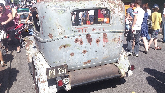 Camberley Town Centre car show - Page 1 - Thames Valley & Surrey - PistonHeads - This image depicts a lively street scene, where the main focus is a vintage car with a distinctive white exterior and a pinkish underbelly. The car has a weathered appearance with noticeable rust and patches of paint missing, giving it a rustic charm. In the backdrop, there are several onlookers, some of whom are engaging in various activities like cycling and walking. The car is parked, and the people appear to be observing it with interest. A small amount of graffiti, including an "X" with "4" underneath it, is visible on the car's hood, adding to its vintage appeal.