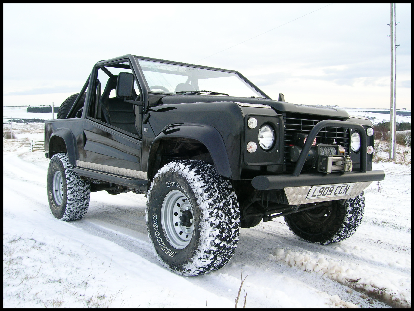 Pistonheads Kit Defender - The image features a rugged black off-road vehicle, likely an SUV, with a bulky frame and large round tires. The truck appears to be equipped with a roll cage, a black roof rack, raised off the ground on its huge tires, and red neon lights. The vehicle is situated on a snowy terrain with small hills and what looks like a pole or structure is barely visible in the distance.