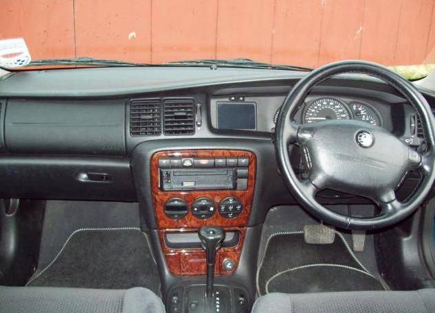 Vectra - Page 1 - Readers' Cars - PistonHeads - The image shows the interior of a car with a focus on the driver's seat area. Notably, the car has a wooden or laminate dashboard, which provides a nice contrast to the black body. On the dashboard, there are several control buttons, indicative of an older model car, possibly a Japanese vehicle due to the round steering wheel. The cabin features a gray upholstery style, and the steering wheel has some Japanese script on it. There is a limited view of the car's exterior visible in the background, featuring a red brick wall. There is also a small round object resembling a speedometer or a gauge of some kind on the dashboard.