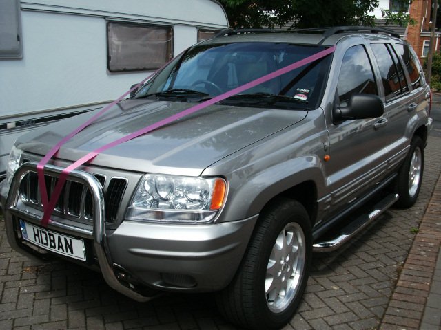 A white truck parked in a parking lot - Pistonheads - The image presents a scene featuring a silver SUV prominently positioned on a brick surface. The vehicle exhibits a sleek aesthetic, its silver paint gleaming under the light. A notable detail on the SUV is the dark purple ribbon that is wrapped neatly around its grille. This ribbon isn't just for decoration, as the word "HONDA" can be discerned on it, suggesting a possible affiliation with the Honda brand. In the background, partially obscured, there is a white house and a trailer, adding to the everyday ambiance of the scene.