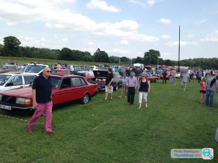 Corbridge classic show - Page 1 - North East - PistonHeads - This image depicts an outdoor scene where several people are walking across a lush green field that is lined with parked cars. The weather appears to be overcast, with the sky filled with clouds. In the foreground, a man dressed in a black shirt and pink pants is walking towards the viewer, looking directly at the camera. In the background, various other individuals can be seen observing the cars and the field. The cars are parked in neat rows, creating a distinct boundary between the grassy field and the parked vehicles. The photo appears to have been taken during the day, likely on a weekend, as indicated by the casual clothing and leisurely activities of the people.