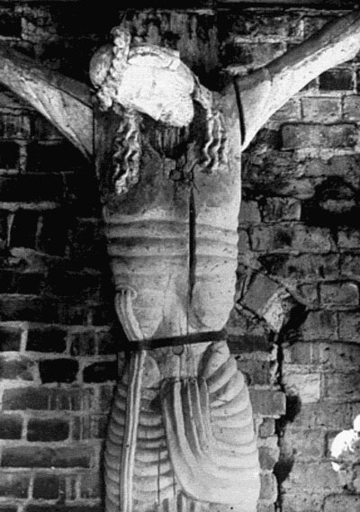 The image appears to be a historical photograph of a rustic, handcrafted figure, possibly of religious significance, mounted against a weathered, brick wall. The figure is dark in color with captured details highlighting muscle definition and intricate patterns. It's a monochrome image, likely from the 20th century, that has an aged appearance with some elements of decay and wear. The binding on the figure's neck and lower back traces a harsh history of customs or rituals once practiced.