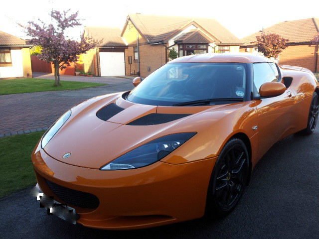 The future is bright.......... - Page 1 - Evora - PistonHeads - The image features a parked orange sports car on a driveway adjacent to a residential area. The car has a sleek design, with a modern front and black wheels. There is a hint of a dark color for the background, which contrasts with the bright orange of the car. The sky appears gray and overcast, suggesting it might be a cloudy day.