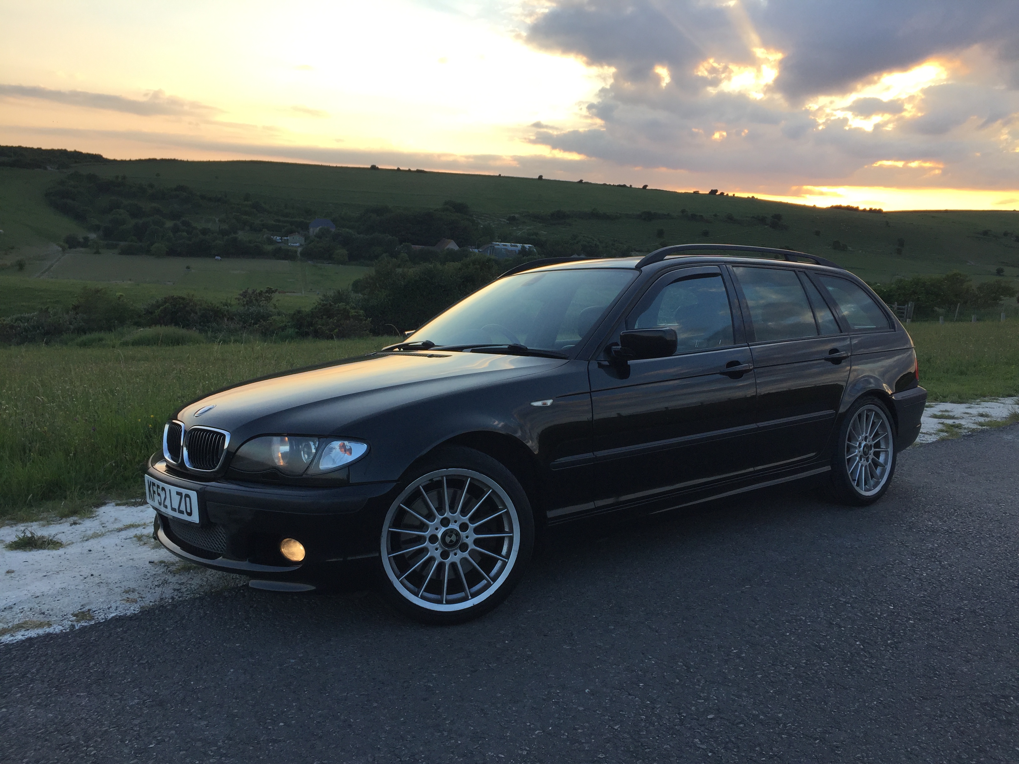 BMW E46 330d M-Sport Touring Manual (Anyone recognise her?) - Page 2 - Readers' Cars - PistonHeads - The image shows a car parked on the side of a road, with a beautiful sunset in the background. The car is black with white wheels and appears to be a BMW. It's positioned at an angle that gives a clear view of its side profile. There are no people visible in the scene. In the distance, there's a house and some greenery, contributing to a serene rural setting.