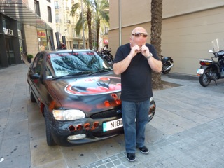 "Chip run cars win in Barcelona" - Page 1 - East Anglia - PistonHeads - The image shows a person standing next to an eccentric, brightly colored, and customized car that resembles a four-wheeled motorcycle with a wide wheelbase and two seats. The individual appears to be a motivational speaker, wearing a black shirt, glasses, and blue jeans, with a microphone in hand, seemingly in the middle of a presentation or tour. The setting appears to be a city street, with palm trees lining the background and other city vehicles and landscaping elements visible in the vicinity of the car.