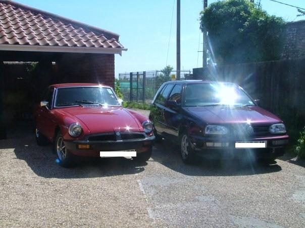 My Cars! - Page 1 - Readers' Cars - PistonHeads - The image shows a driveway with two vintage cars parked side by side. One car is red and the other is maroon. Both vehicles have a shiny finish reflecting light, possibly suggesting they are well maintained. The background features a solid, non-descript wall. The overall scene suggests a suburban setting.