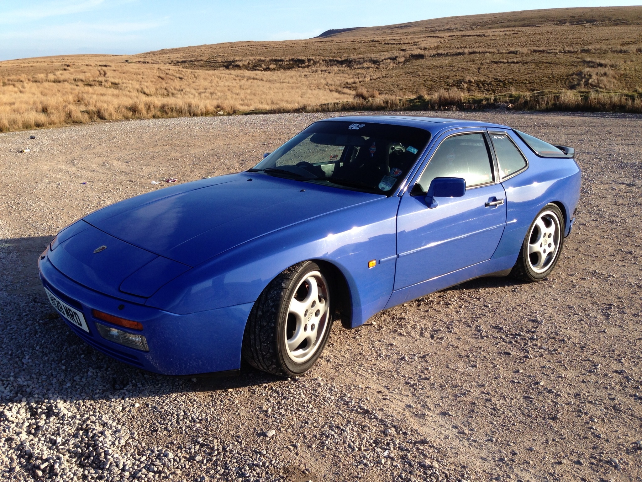 Picture your Porsche - Page 1 - Front Engined Porsches - PistonHeads - The image showcases a vibrant blue Porsche Cayman, which is parked on a gravel field. The setting sun casts a warm glow on the vehicle and the surrounding landscape. In the backdrop, rolling hills extend into the horizon, and the sky is painted with hues of sunset. The car's polished exterior contrasts with the rugged terrain, adding a sense of luxury to the rustic environment.