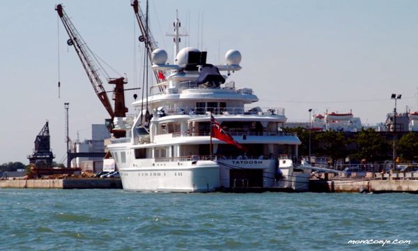 Monacoyachts - The image shows a large luxury yacht motorboat that is tied up to a dock. The boat is a Tahitosh owned by a person named Mosaad. There are three flags, possibly of the vessel's origin, flying on poles on the ship. There are several people on deck, who appear to be passengers or crew members. The boat is moored near a large construction crane, indicating that the area might be under development. The backdrop features a clear blue sky, hinting at fair weather.