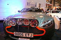 Pistonheads - The image features a contemporary small car at an indoor or outdoor event. The vehicle has a sleek, aerodynamic design with orange and gray detailing, prominent headlights with a circular, arranged pattern, and a license plate that reads "O006 ETJ". The car is on display, with lights reflecting on the side of the vehicle. In the background, there are other cars, suggesting this could be a car show or exhibition.