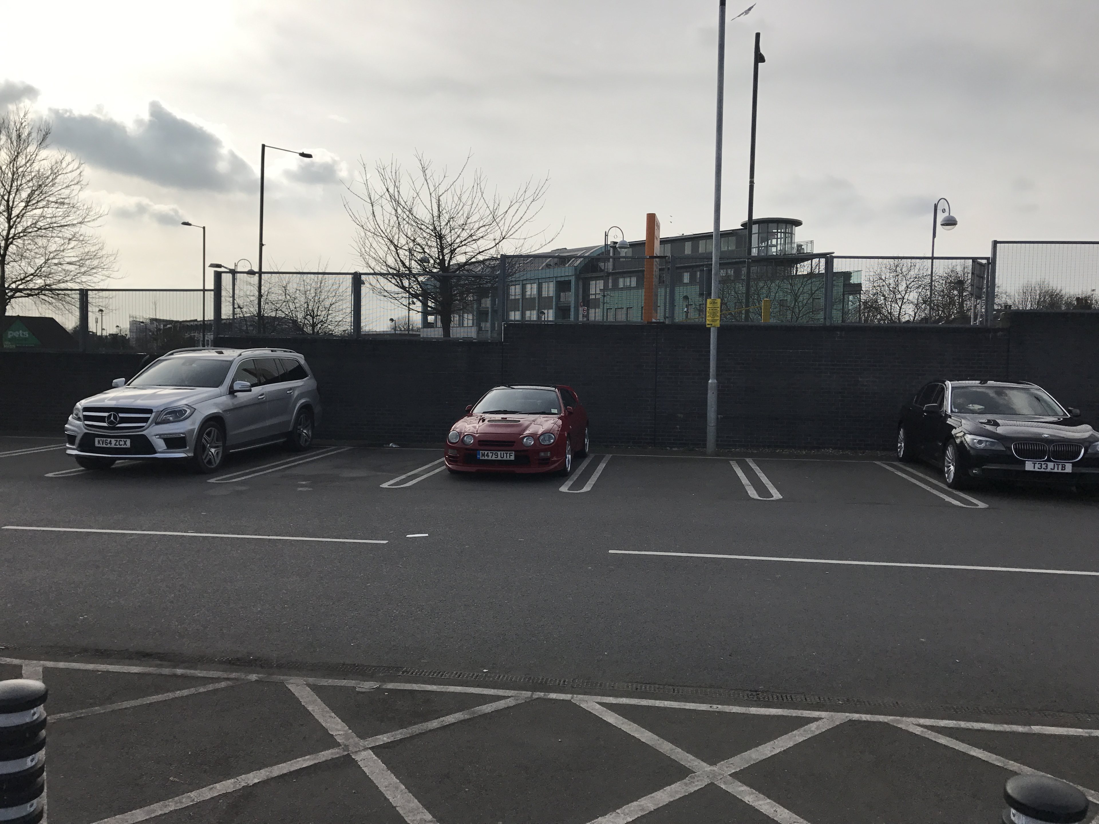 Classics dwarfed by moderns - Page 130 - Classic Cars and Yesterday's Heroes - PistonHeads UK - The image is a photograph of an outdoor parking lot. There are several cars parked, including one that appears to be a sports car with a red hue. In the background, there's a building and a street sign with the number 31 on it. A fence is visible, separating the parking area from another space or property. The sky is overcast with clouds, suggesting an overcast day. There are no people immediately visible in the scene.