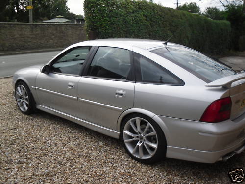 Gsi Pistonheads Vectra - The image shows a silver sedan parked on a gravel driveway. The car is positioned at an angle, slightly tilted to the left, providing a clear view of its sleek design and chrome wheels. The sedan has small tinted windows and a sunroof, adding to its sporty appeal. A neatly folded flag is visible on the back, possibly indicating a country or team affiliation. The driveway on which the car is parked is lined by a brick wall and neatly trimmed hedges, contributing to the overall pleasant and well-maintained appearance of the scene.