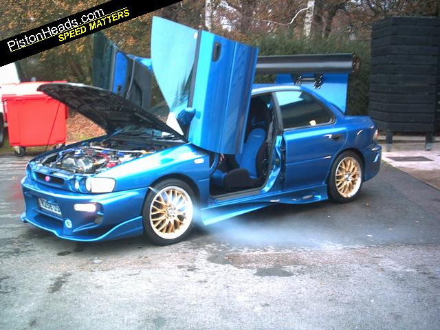 Badly modified cars thread - Page 46 - General Gassing - PistonHeads - The image depicts a blue car parked in a lot. The car is open, with the hood up, revealing a well-maintained engine under the hood. The car appears to be a converted sports car, as suggested by the visible modification elements and the excited expression of the image's text, proclaiming it as the "PistonHeads Speed Matter." The car's wheels are fitted with a distinctive multi-spoke design. The setting includes a few trees in the background, indicating an outdoor parking area.