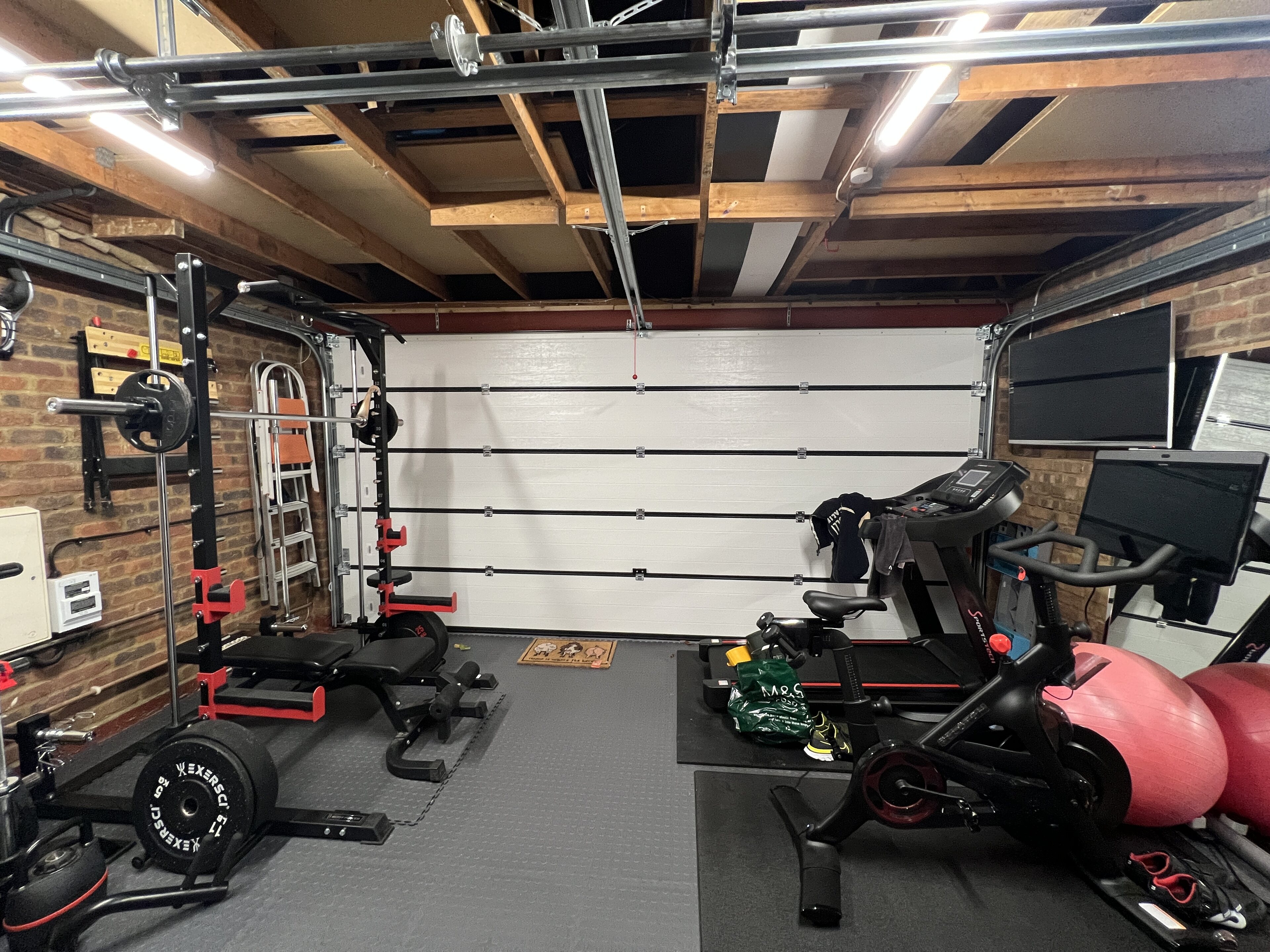 double width garage door conversion - Page 2 - Homes, Gardens and DIY - PistonHeads UK - The image shows a well-equipped home gym. Dominating the center of the room is a large workout machine, which seems to be a treadmill or elliptical trainer given its elongated structure and footrests. To the right of this central piece, there's a weight bench with various weights and possibly an exercise ball placed nearby. The wall behind the equipment features a brick-like texture, adding a touch of rustic charm to the room. A large mirror is mounted on the wall opposite the machine, reflecting the room and its contents. The floor is covered in a rubberized mat, typical for gyms to provide cushioning and prevent slipping during workouts. Despite the presence of weights and other equipment, there's a sense of orderliness and cleanliness in the space, suggesting it's well-maintained and cared for.