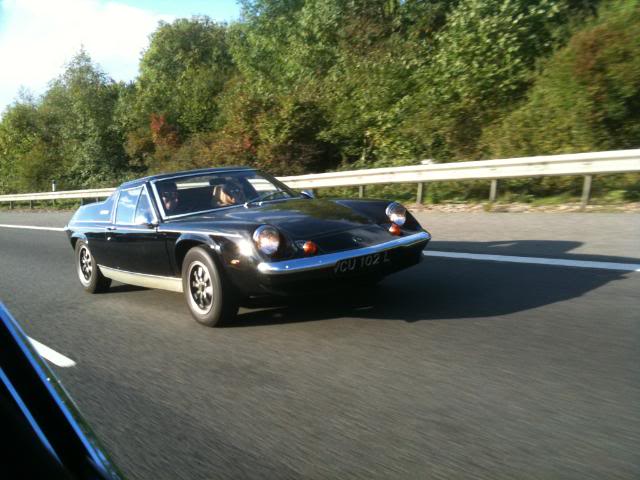 Reliable Shop Repair Special Pistonheads Lotus Europa - The image shows a vintage car driving on a highway. The car is dark and has headlights that are currently on. It features two round lights, possibly front turn signals, on each side of the vehicle. A driver is visible in the front seat who appears to be operating the car. The background indicates a natural setting with lush green trees and what appears to be a guardrail on the side of the road. The road itself is paved and curves to the left ahead of the car, with a solid white line marking one side of the road. There are no visible texts or distinctive marks indicating the car's make or model.