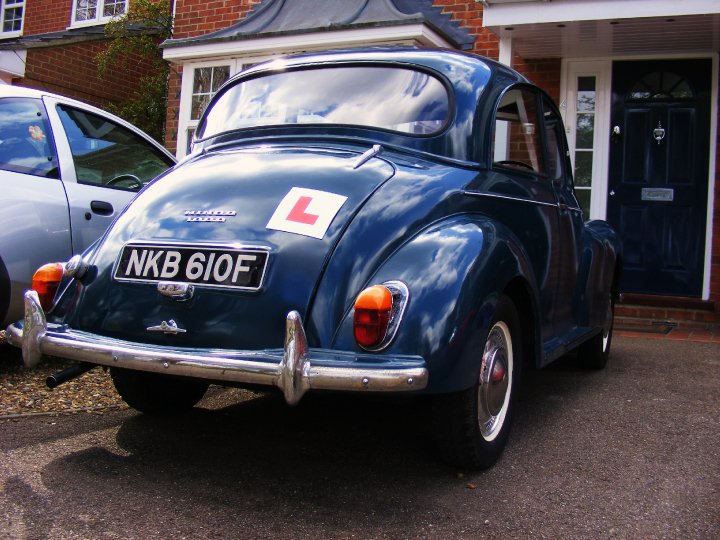 Shopping Pistonheads - This image features a classic vintage vehicle painted in a deep shade of blue. The car is positioned on a driveway against the backdrop of a house with a brick facade. A red "L" sign is prominently displayed on the trunk, and the license plate reads "NKB 61O." The vehicle has white walls on the tires that add to its vintage appeal. The setting suggests a residential area, possibly in an urban neighborhood.