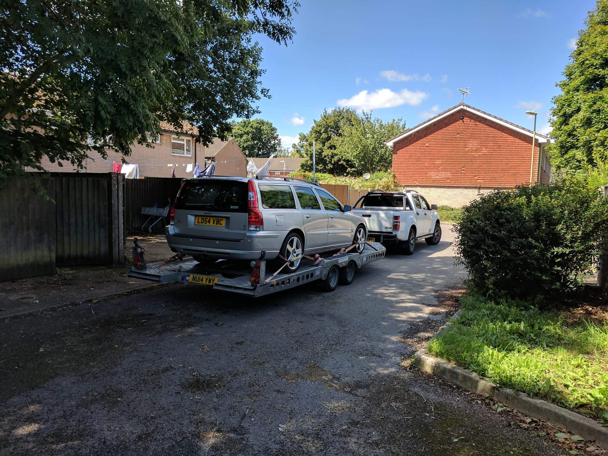 154k Volvo V70....R you feeling brave? - Page 11 - Readers' Cars - PistonHeads - The image depicts a tune-up road trip scenario. It features two cars and two tunes-up cars, one white pickup truck and one white sedan. They are parked in front of a wooden barn and a tree. The pickup truck is being pulled by a flatbed trailer, and it has a red flag attached. There is also an additional white car at the back of the flatbed trailer. The image has a clear daytime setting with ample natural light.