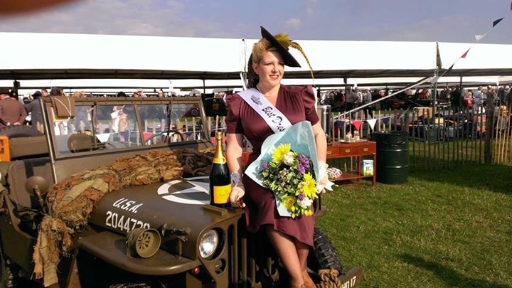 A woman standing next to a luggage cart - Pistonheads - The image depicts a woman with light brown hair, dressed in a red dress and a maroon hat adorned with a feather. She is resting an arm on an antique military jeep, which has a classic World War II style. The vehicle is decorated with a snake skin pattern on its side, and the front adorns a label reading "USA-AT1.M.G.INPH." The overall scene is set outdoors, as the woman, dressed as a queen, is surrounded by pieces of vintage military equipment. There are additional elements, like a champagne bottle and a presentation box of flowers, suggesting a festive or celebratory occasion.